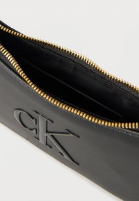 Μαύρο δερμάτινο πουγκί με εγχάρακτο λογότυπο CK, χρυστό μεταλλικό φερμουάρ μισάνοιχτο, που αποκαλύπτει μαύρη υφασμάτινη επένδυση εσωτερικά.