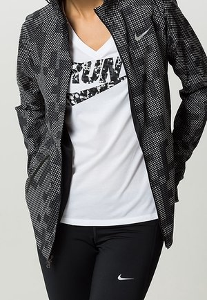 Osoba nosząca czarno-białą kropkowaną kurtkę na białej koszuli z tekstem "RUN" oraz czarne spodnie z białym logo Nike.
