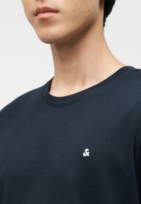 Κοντινό πλάνο ενός ατόμου που φοράει μπλουζάκι με στρογγυλή λαιμόκοψη σε σκούρο μπλε χρώμα, με ένα μικρό λευκό σύμβολο Άνπερσαντ αριστερά στο στήθος.