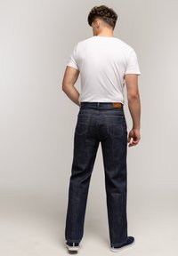 Mężczyzna stojący tyłem, ubrany w białą koszulkę, ciemnoniebieskie dżinsy o prostym kroju i granatowe trampki, na jednolitym jasnym tle.