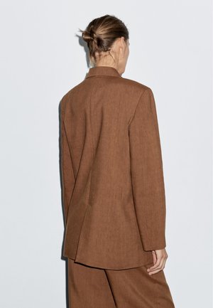 Brauner Leinenblazer mit maßgeschneidertem Design, einfacher Rückenbelüftung und langen Ärmeln. Schlichte, klare Linien. Getragen über passenden Hosen.