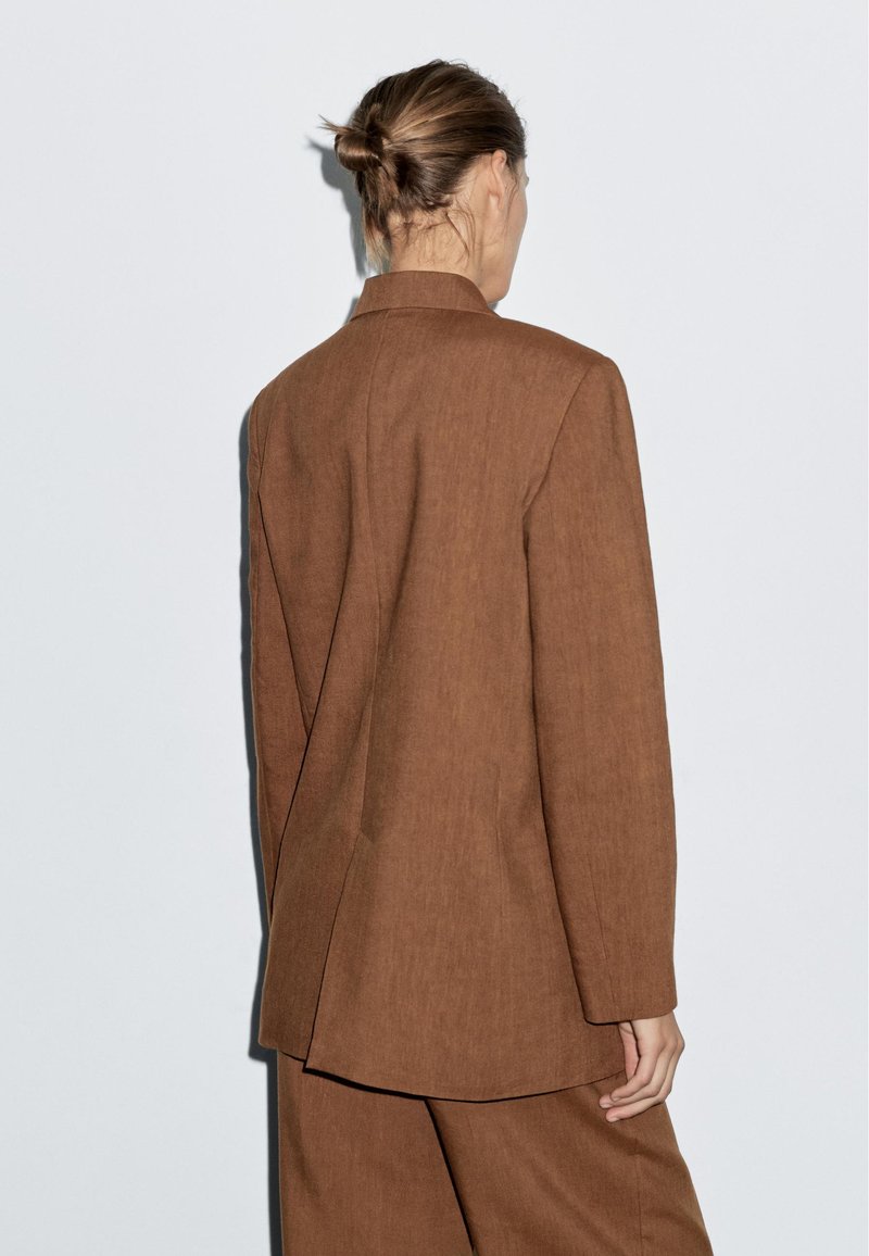 Blazer en lin brun avec un design ajusté, une fente au dos, et des manches longues. Lignes simples et épurées. Porté sur un pantalon assorti.