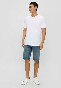 T-shirt en coton blanc à manches courtes, associé à un short en denim bleu clair. Le modèle porte des baskets blanches, se tenant devant un fond neutre.