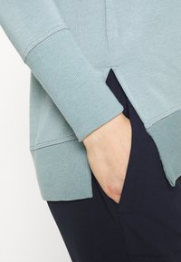 Helles blaues Strickpullover mit gerippten Bündchen und seitlichen Schlitzen. Über dunkelblauen Hosen getragen, die einen Kontrast in der Stoffstruktur zeigen.