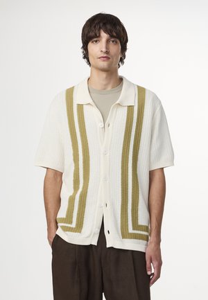 Cardigan à col et manches courtes en crème avec des rayures verticales vertes. Présente une texture tricotée et une fermeture à boutons sur le devant.