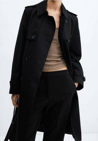 Schwarzer Trenchcoat mit doppelt geknöpftem Design, großen Knöpfen und Schulterklappen. Getragen über einem hellbraunen Oberteil und schwarzen Hosen.