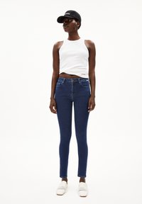 Jeans skinny bleus avec une texture lisse, associés à un débardeur blanc à côtes. Le modèle porte une casquette noire et des chaussures slip-on blanches en tissu.