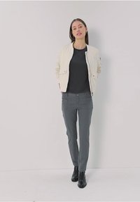 Veste bomber crème clair sur une chemise noire, associée à un pantalon slim gris et des bottines noires. Texture lisse et design ajusté.