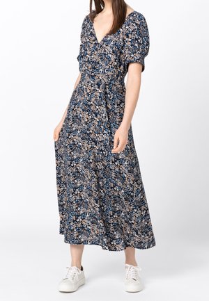 Sukienka kopertowa w kwiaty z granatowym tłem i jasnym kwiatowym wzorem, krótkimi bufkami, dekoltem w kształcie litery V i długością do połowy łydki, w zestawieniu z białymi trampkami.