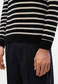 Suéter a rayas negro y crema hecho de tejido de punto con un dobladillo acanalado. Presenta un sutil logo "MN" en el costado. Combinado con pantalones oscuros.