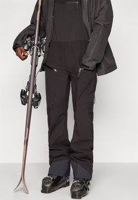 Juodi slidinėjimo kelnių su vandeniui atspariu paviršiumi, siaurėjančiomis kojomis, šoninėmis užtrauktukais ir sustiprintomis sijonų dalimis. Derinama su slidinėjimo batais ir slidėmis.