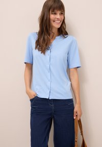 Helles blaues kurzärmliges Hemd mit Rundhalsausschnitt, lockerer Passform und glatter Textur, kombiniert mit dunklen Jeans.