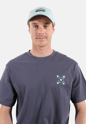 Jeune homme portant une casquette Oxbow bleu clair et un t-shirt gris foncé avec un petit motif floral sur la poitrine, faisant face à l'avant sur un fond uni.