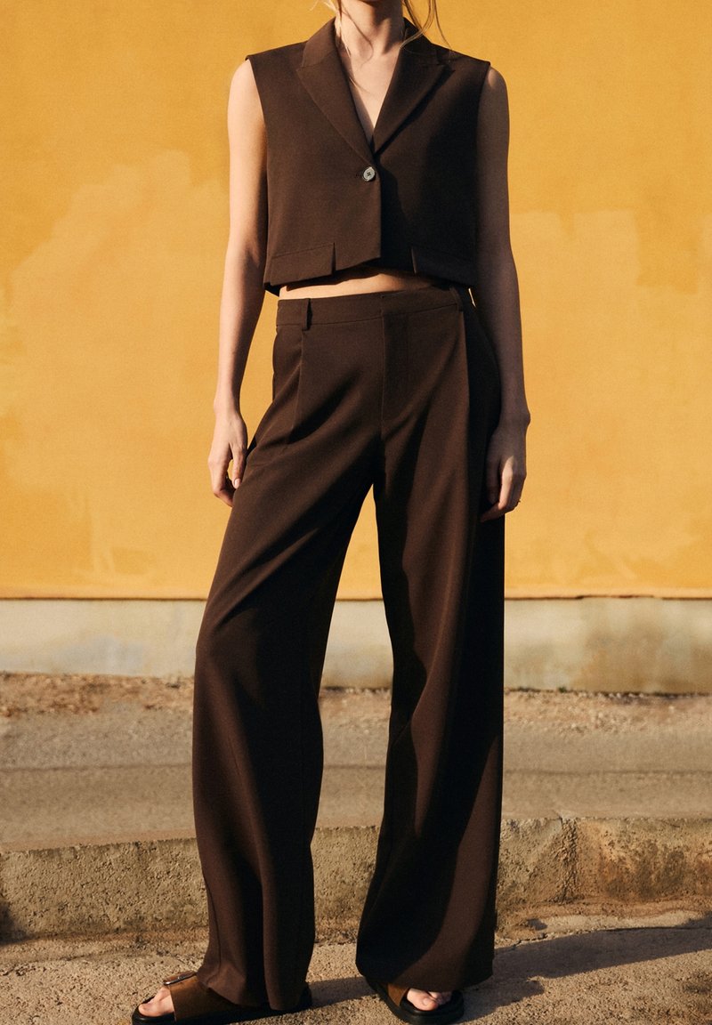 Femme portant un blazer court sans manches marron foncé et un pantalon à jambes larges, debout contre un mur jaune en plein soleil.