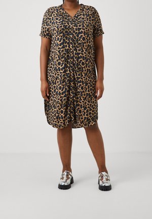 Person iført en knælængde kortærmet kjole med leopardprint og sølvfarvede sko med chunky sål, stående mod en ensfarvet baggrund.