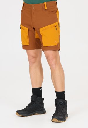 Mann trägt braune und gelbe Wandershorts mit großen Reißverschlusstaschen, schwarze Socken und schwarze Wanderschuhe vor weißem Hintergrund.