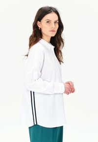 Camisa blanca de botones con mangas largas, que presenta un dobladillo curvado y rayas laterales negras. Tejido suave con un diseño clásico de cuello.