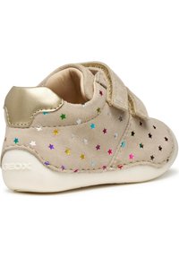 Scarpa in suede beige con accenti dorati metallici e motivi a stella multicolori. Presenta una fascia in velcro e una suola in gomma bianca.