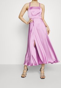 Robe en satin rose longue avec un décolleté carré, des fines bretelles et une fente sur le côté. Associée à des talons hauts argentés à lanières.