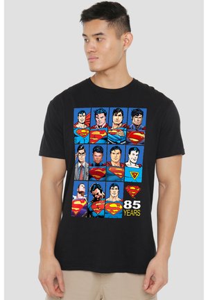 Mann trägt ein schwarzes T-Shirt mit einem Raster von zwölf Superman-Comicstilen und dem Text "85 years" sowie dem Superman-Logo unten.