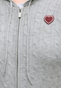 Sweat à capuche zippé gris clair en maille torsadée avec cordons de serrage et petit écusson cœur rouge sur le côté gauche de la poitrine.