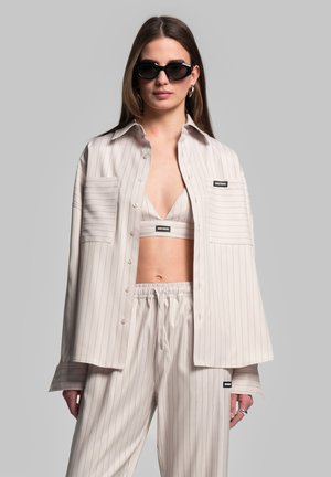 Light beige pinstriped shirt with two chest pockets, worn over a matching crop top and relaxed-fit pants, paired with sleek black sunglasses.