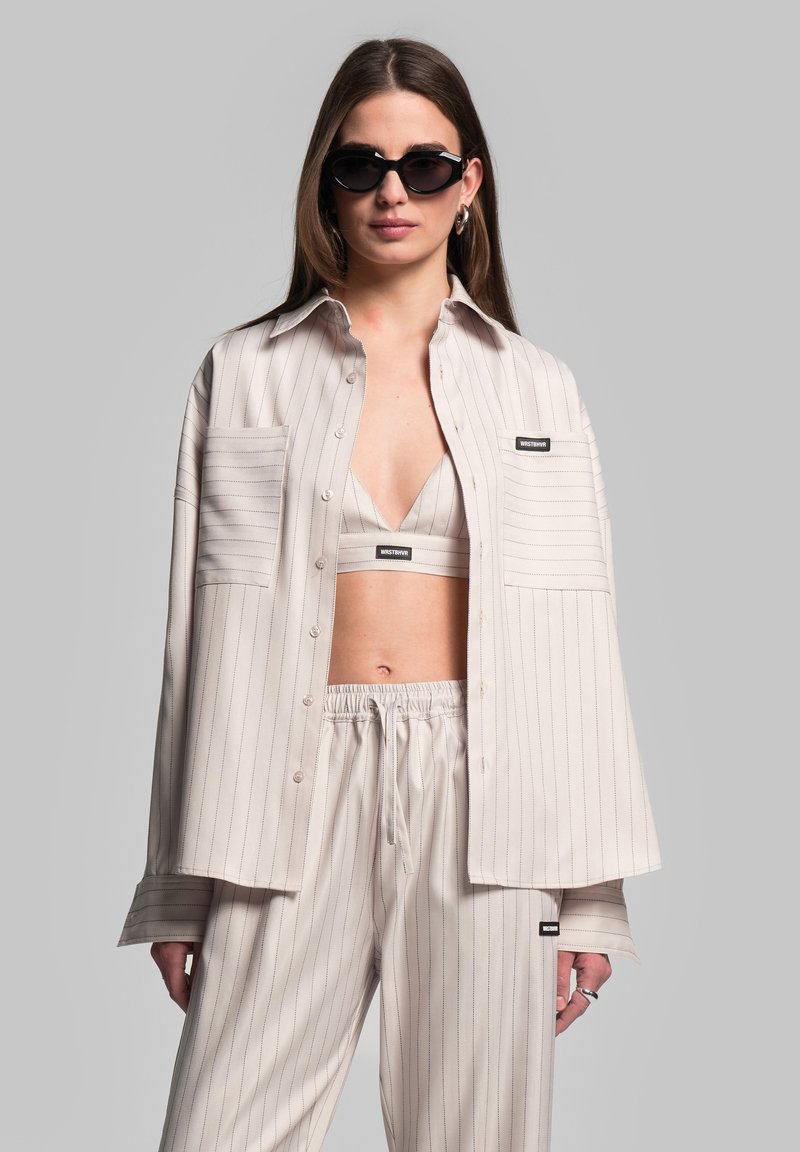 Lichtbeige gestreepte blouse met twee borstzakken, gedragen over een bijpassende crop top en een ontspannen broek, gecombineerd met stijlvolle zwarte zonnebril.