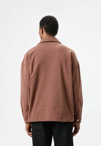 Bruine fleecejas met een losse pasvorm, grote kraag en omgeslagen mouwen. Soepele textuur met strakke lijnen en minimale accenten.