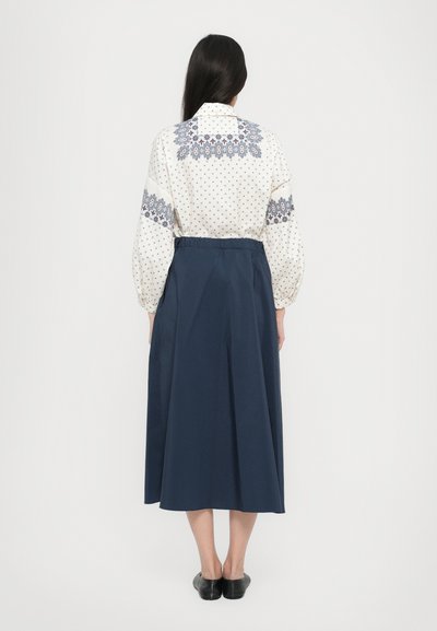 Langärmlige Bluse in Creme mit blauer, gemusterter Stickerei, kombiniert mit einem dunkelblauen, fließenden Rock. Schwarze Schuhe runden das Outfit ab.