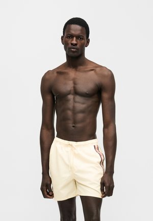 Homme noir athlétique debout, torse nu, portant un short de bain crème avec des bandes latérales, sur un fond blanc uni.