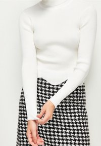 Haut blanc à manches longues côtelées avec une silhouette ajustée et un ourlet festonné, associé à une jupe à motif pied-de-poule noir et blanc.