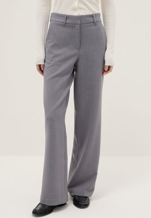 Donna con pantaloni grigi a righe a gamba larga, maglia bianca a maniche lunghe in tessuto a coste e mocassini neri, in piedi su sfondo bianco.