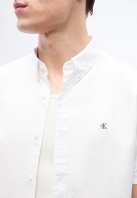 Νέος άνδρας που φοράει λευκό πουκάμισο με κουμπιά και μικρό λογότυπο "CK" στο στήθος πάνω από λευκό ριγωτό φανελένιο εσώρουχο, κοιτάζει προς τα αριστερά, με ορατό λαιμό και κάτω μέρος προσώπου.