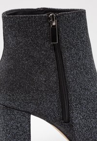 Svart ankelboot med en texturerad, glittrig yta. Har en sidoglider och en åtsittande design som en strumpa. Hög klack för extra höjd.