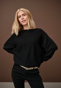 Szőke nő fekete pulóverben és nadrágban, mintás övvel, magabiztosan áll egyszínű barna háttér előtt.