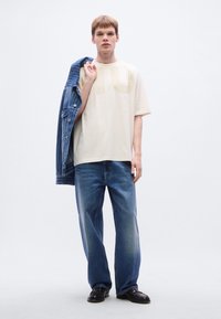 Κρεμ κοντομάνικη μπλούζα με μεγάλα ανοιχτό μπεζ γράμματα, σε συνδυασμό με τζιν παντελόνι με φαρδύ πόδι και μπλε ριγέ τζιν μπουφάν που ακουμπάει σε έναν ώμο.