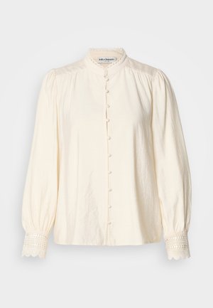 Camicia bianca con colletto alto, chiusura anteriore con bottoni e maniche a sbuffo. Presenta un bordo in pizzo sui polsini e un tessuto leggermente testurizzato.