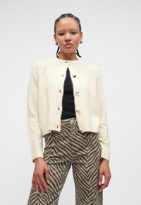 Cardigán krémovej farby z mäkkého materiálu s okrúhlym výstrihom, dlhými rukávmi a gombíkmi v tvare srdca. Kombinovaný so svetrom s pásikmi.