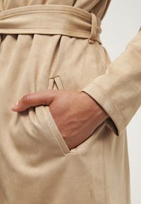 Beige mockaskinnrock med bälte, som visar en sidoficka och en hand som vilar inuti. Texturen ser jämn ut, med subtila veck i tyget.