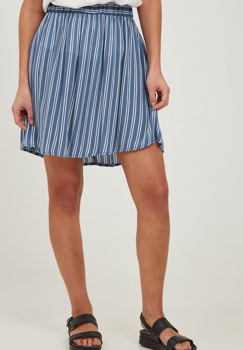 Jupe rayée verticale bleue et blanche avec ceinture élastique portée avec des sandales noires ouvertes au bout, sur une personne debout devant un fond uni.