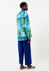 Blaue und grüne geometrisch gemusterte Hemdbluse, kombiniert mit strukturierten blauen weit geschnittenen Hosen und leichten Sandalen.