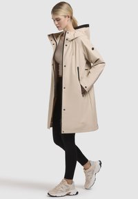 Beige waterdichte jas met een capuchon en zwarte accenten. Voorzien van drukknoopjes, ritszakken en een middenlange uitstraling, gecombineerd met zwarte leggings.