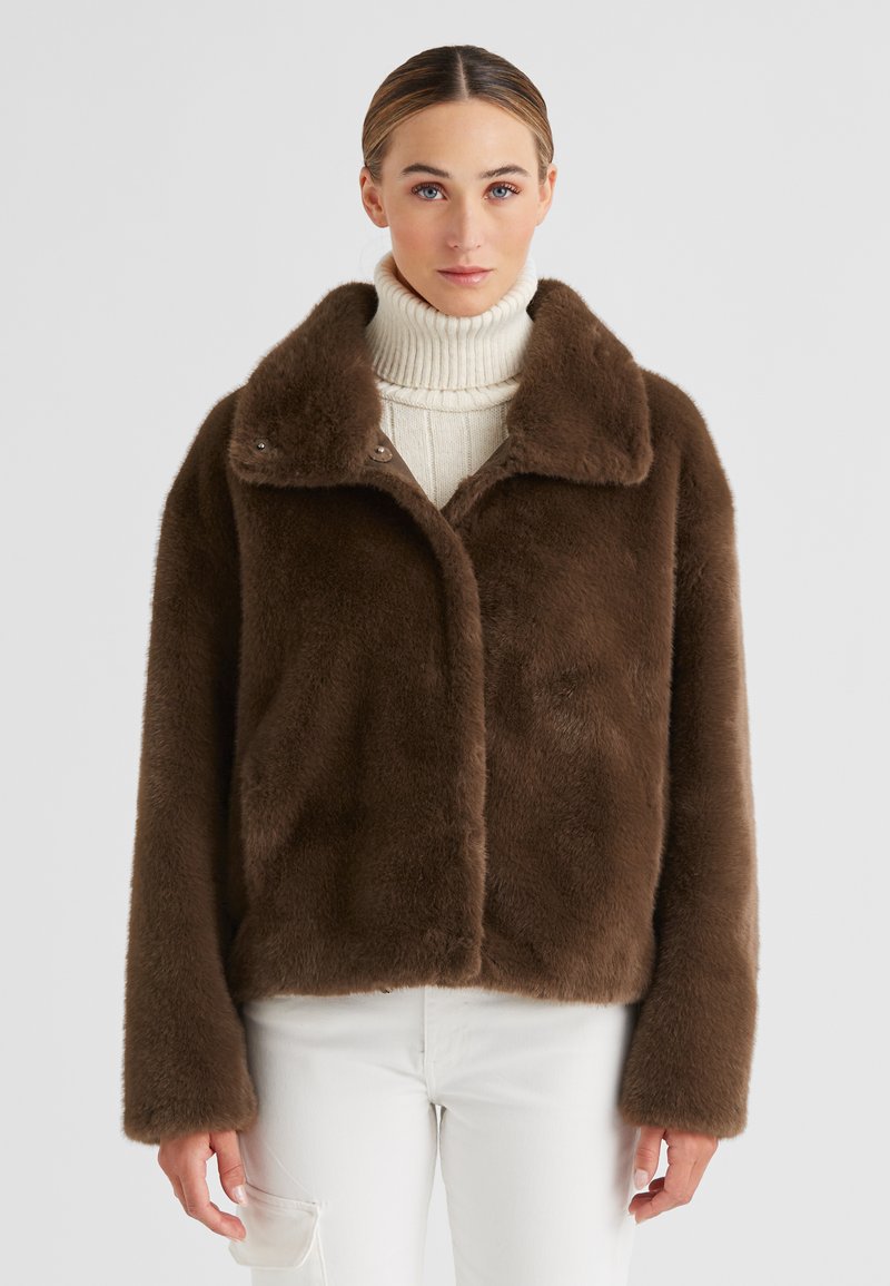 Braune Kunstpelzjacke mit breitem Kragen, weicher Textur und Knopfverschluss, kombiniert mit einem cremefarbenen Rollkragenpullover und weißen Hosen.