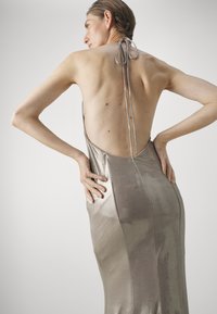 Metallinen halterneck-mekko, jossa on sulavalinjainen siluetti; siinä on syvä selkäosa ja virtaavaa kangasta vaaleanbeige sävyssä.