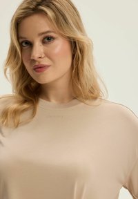 Beżowa bluzka z krótkim rękawem, okrągłym dekoltem i subtelnym logo na piersi. Gładka tkanina i luźny krój, odpowiednia na codzienne noszenie.