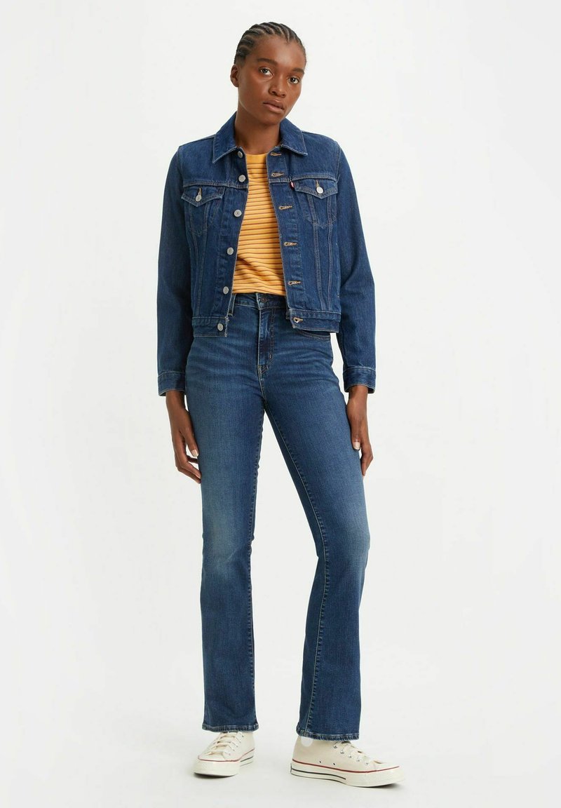 Giacca di jeans in blu scuro, con chiusura frontale a bottoni e due tasche sul petto, abbinata a jeans a vita alta svasati e sneakers bianche.