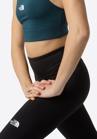 Soutien-gorge de sport cropped en teal et leggings noirs taille haute, avec un logo sur la cuisse, tissu lisse et design ajusté.