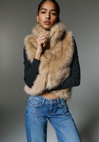 Beige színű műszőrme mellény, magas gallérral és cipzáras záródással. Fekete hosszú ujjú pólóval és kék farmernadrággal viselve.