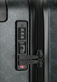 Superficie di una valigia rigida nera con finitura testurizzata, doppie cerniere nere con etichetta "JOOP!", serratura TSA e disco combinatore per la sicurezza.