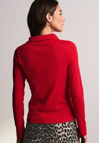 Rød langærmet polo-shirt med ribbet tekstur og krave. Sammen med bukser i leopardprint, der viser kontrasterende mønstre.